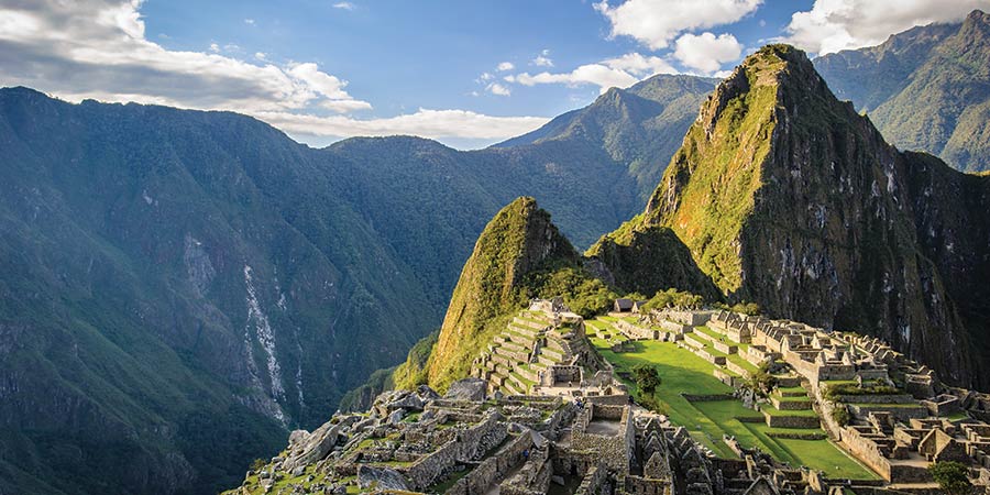 View of Machu Picchu, an ancient stone city built on a mountain. The site is surrounded by green hills and tall mountains under a blue sky with clouds. 
